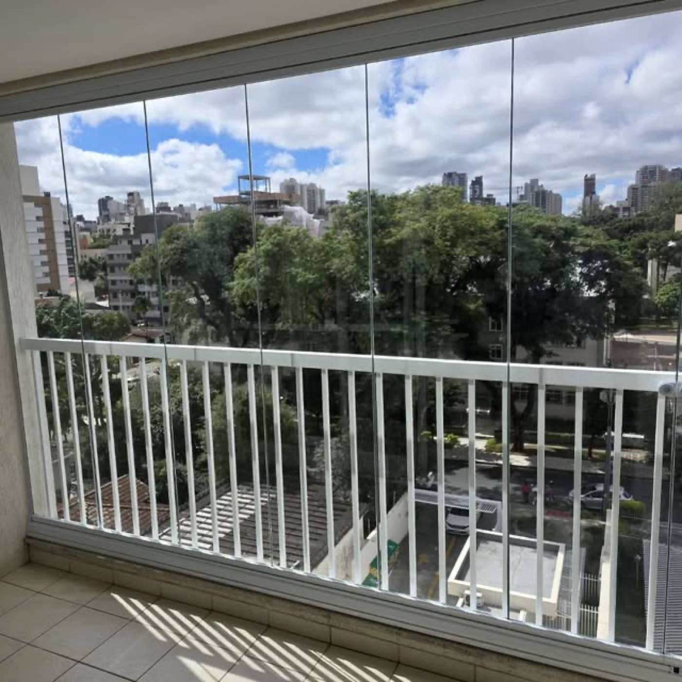 Sacada envidraçada com perfil branco, vista para árvores e prédios em Curitiba